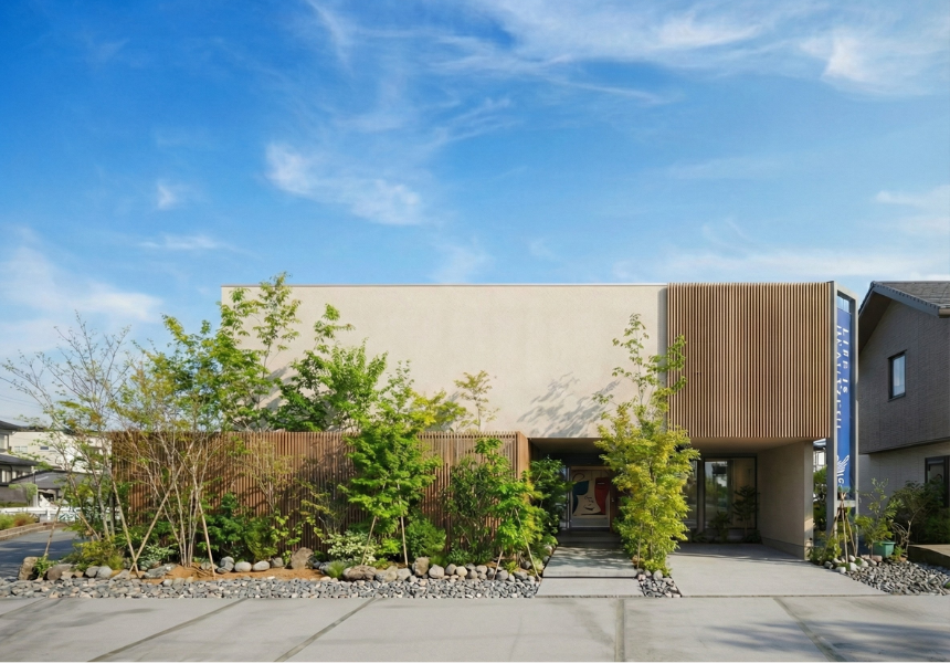 青空に映えるベージュの塗り壁と木製ルーバーが特徴的な和モダンな住宅外観。豊かな植栽が彩るアプローチと、プライバシーを守る洗練されたファサードデザイン。