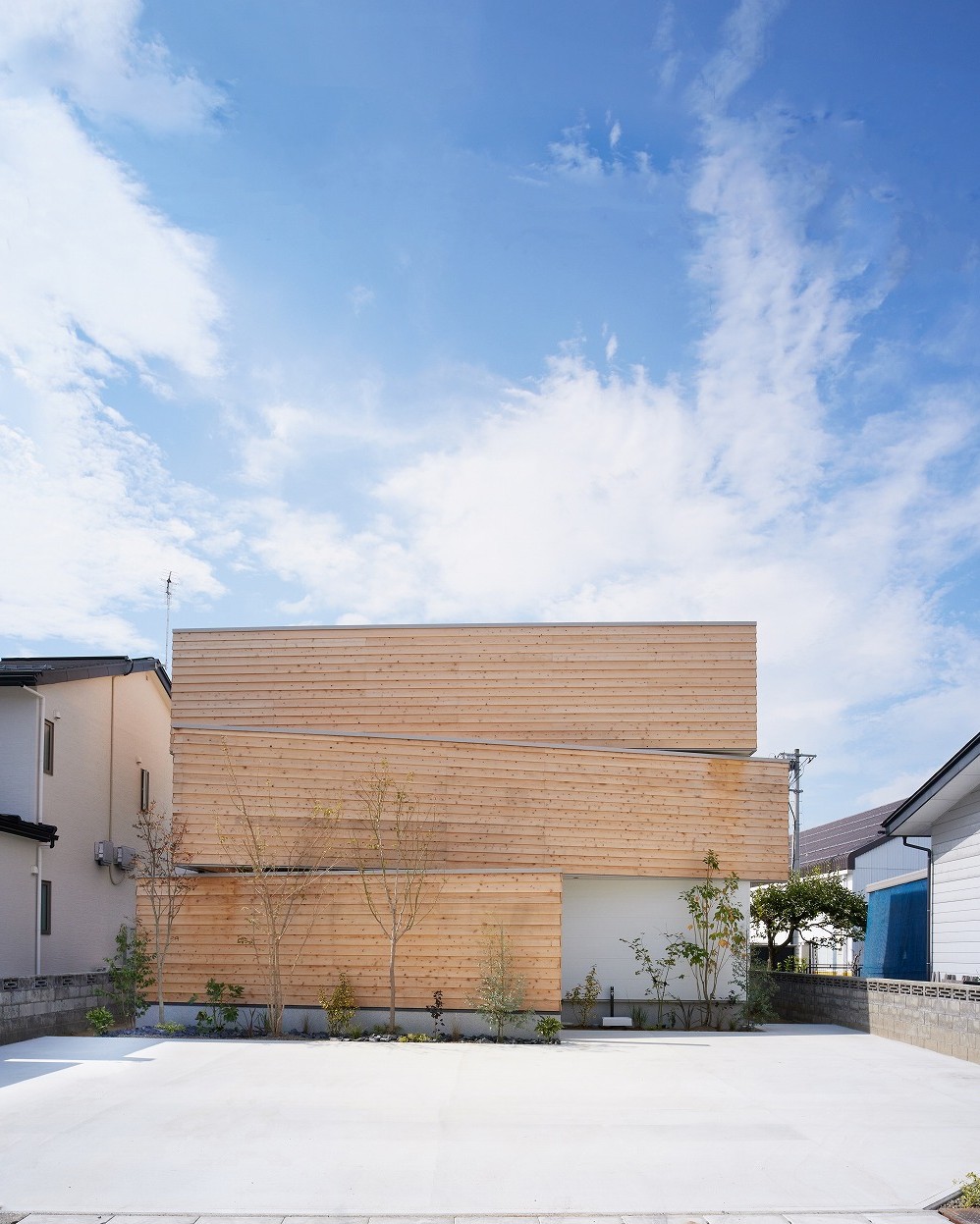 青空の下、木材の外壁が特徴的なモダンな住宅。幾何学的な構造と自然素材のコントラストが美しい。前庭には若木が植えられ、周囲の住宅地に溶け込んでいる。