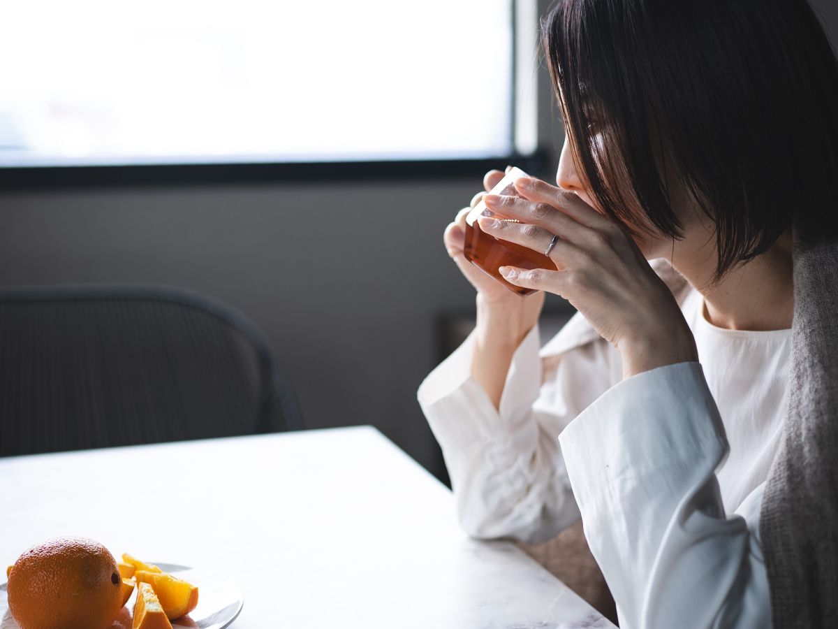 窓辺の明るいテーブルで、白い服を着た女性がグラスでお茶を飲みリラックスしている様子。手前にはカットされたオレンジが置かれている。